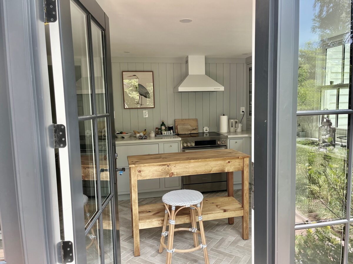A breezy cottage kitchen viewed through French doors, centered on a rustic butcher-block island.
