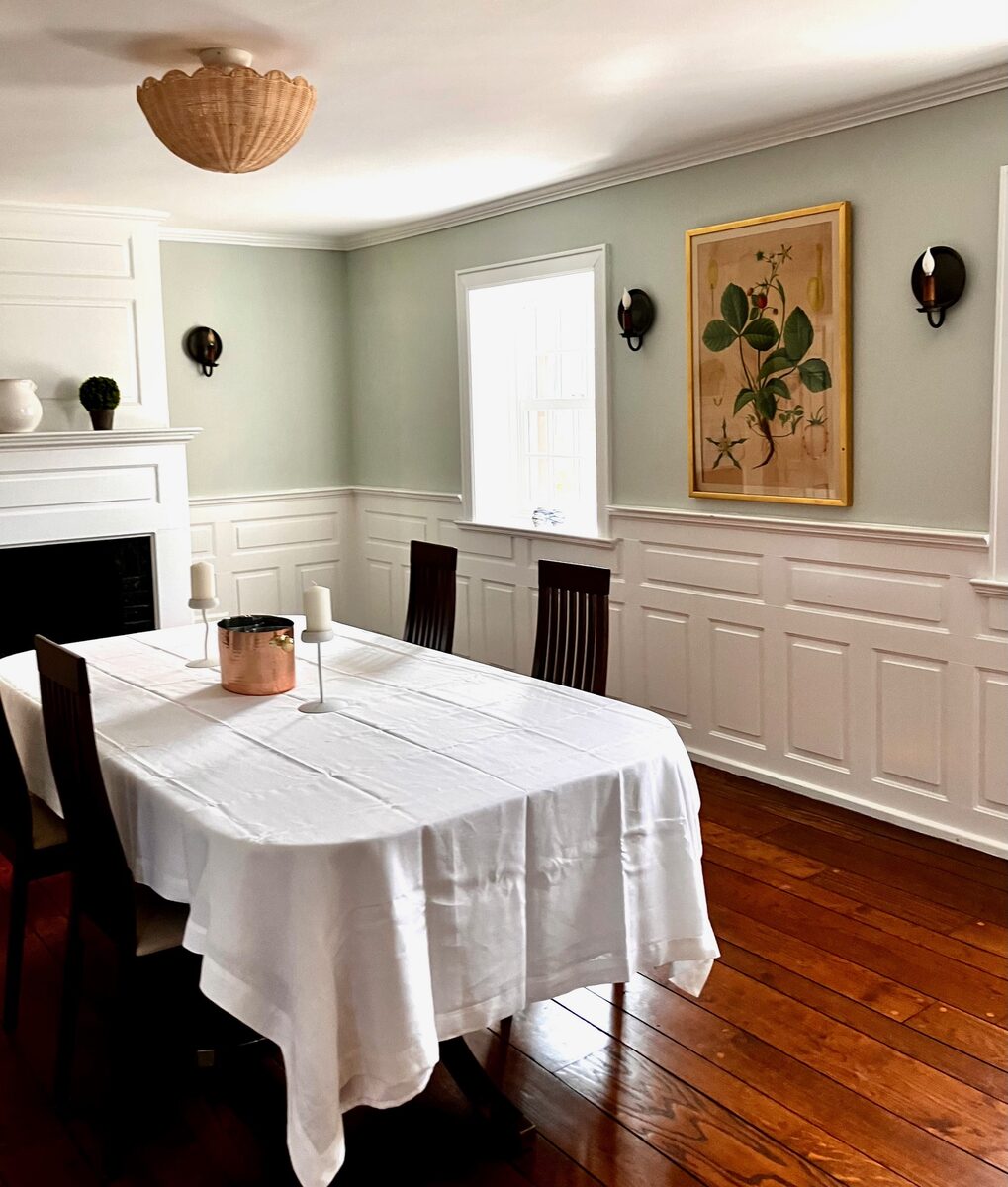 A sage-walled dining room with crisp wainscoting, a single framed botanical, and a shell pendant.