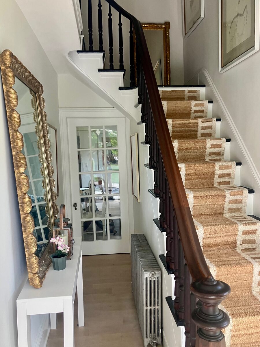 A bright foyer with a banded jute stair runner, a gilt mirror, and a slim console below.
