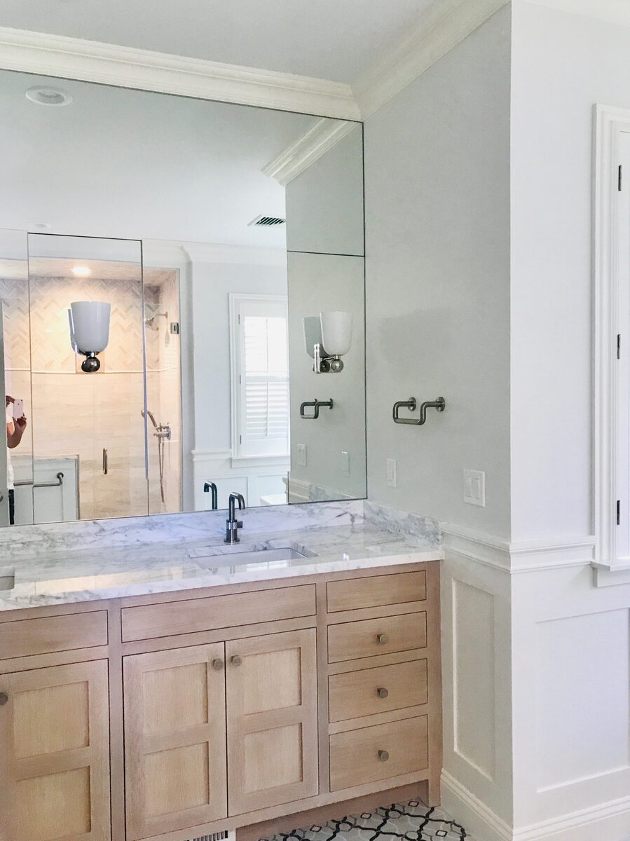 A white oak vanity beneath a frameless mirror, with marble counters and a glass-enclosed shower beyond.