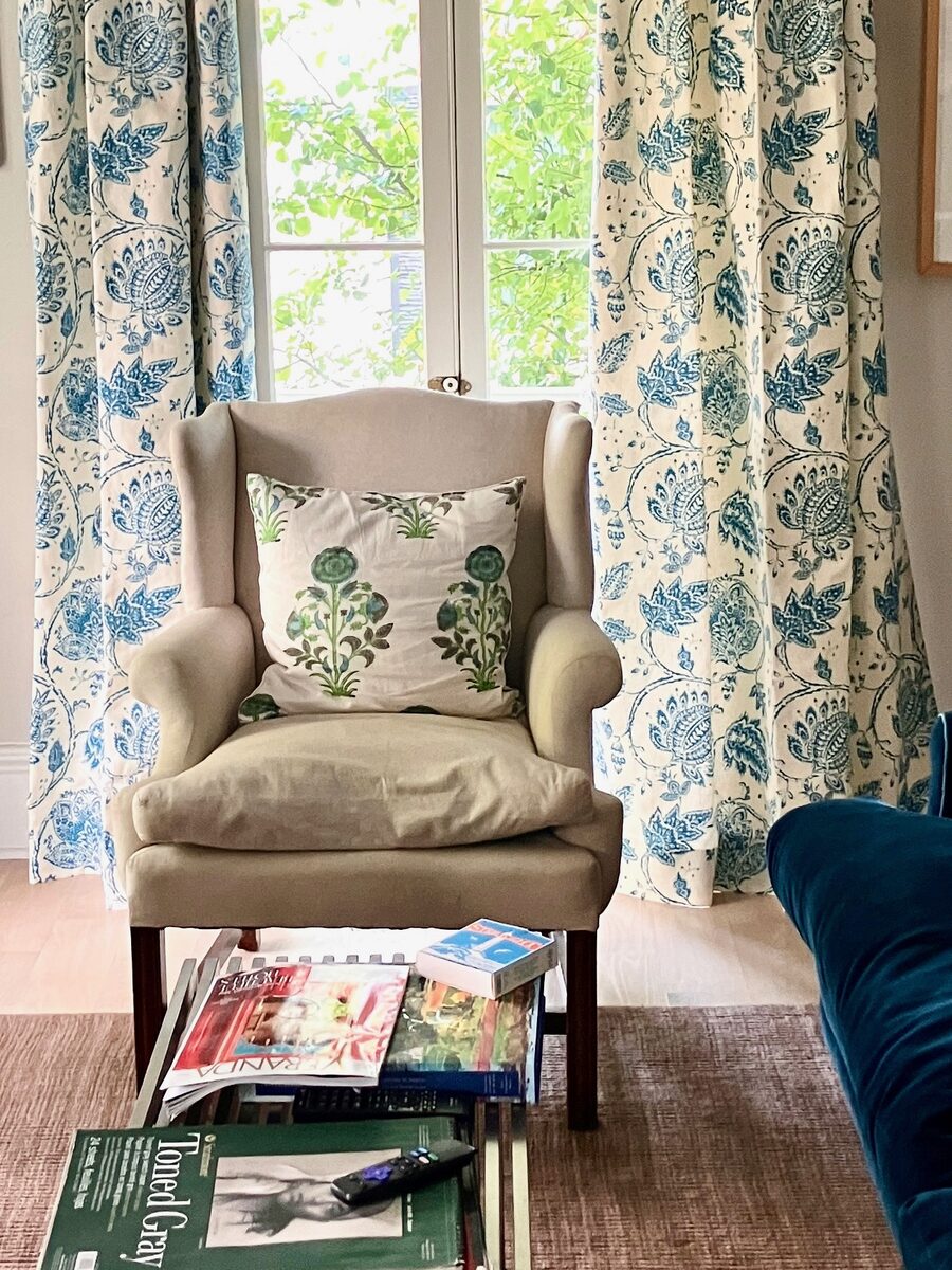 A linen wingback chair framed by blue botanical drapery, with a velvet sofa just in view.