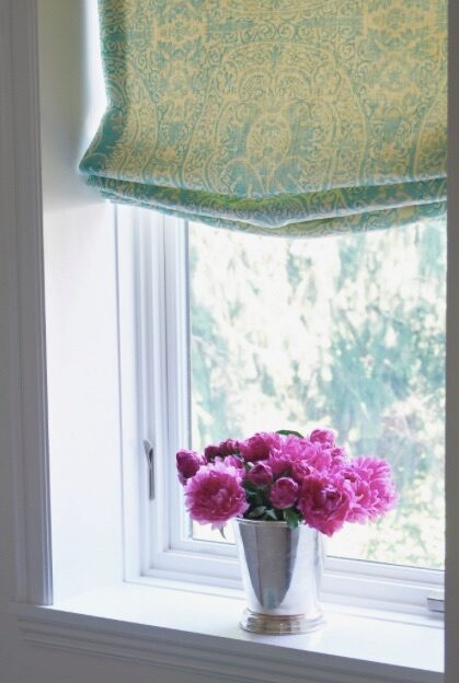 A quiet window vignette with a teal Roman shade and a silver cup of bright pink peonies.