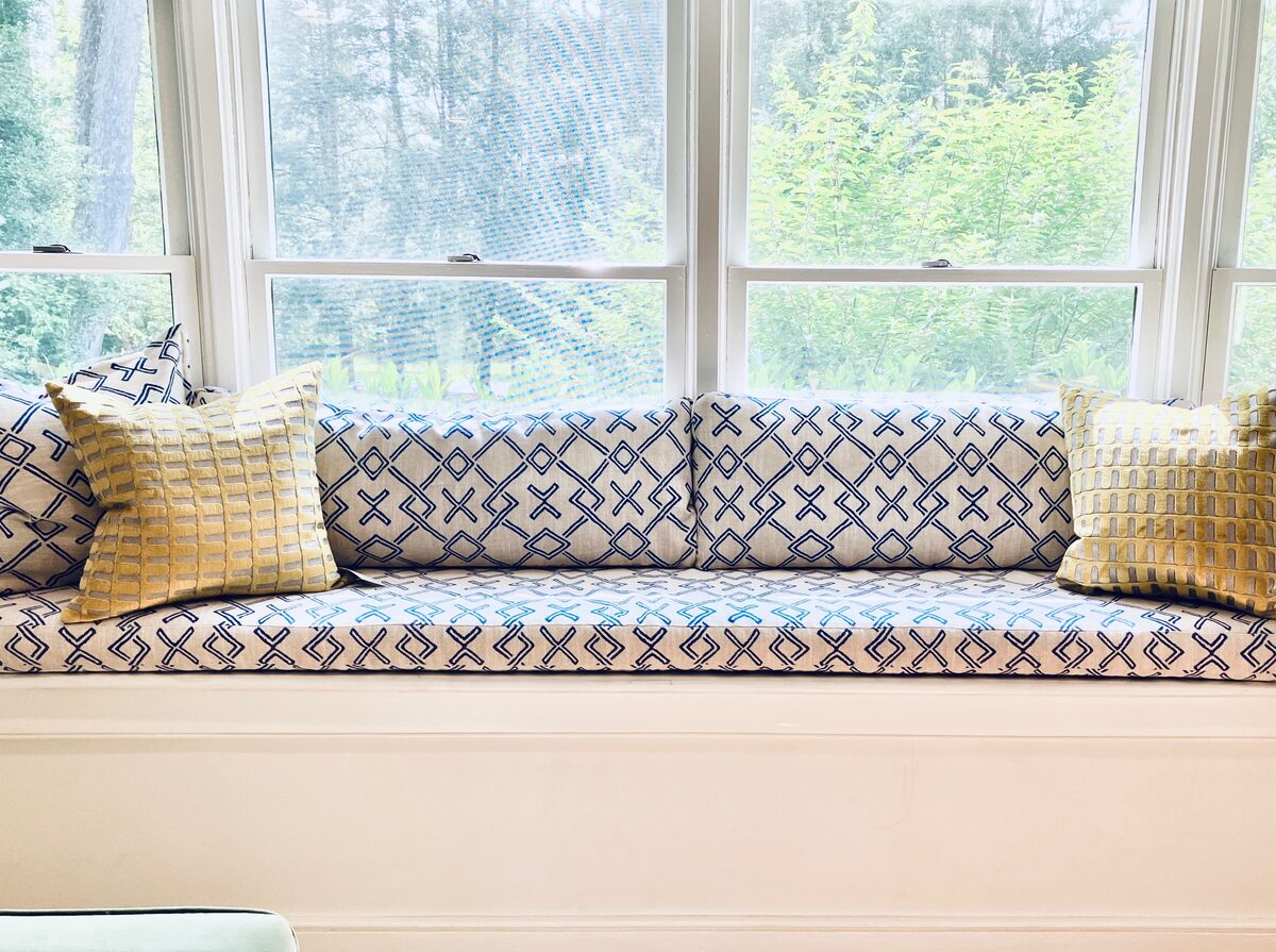 A built-in window seat layered with blue geometric cushions, framed by patterned drapery and garden light.